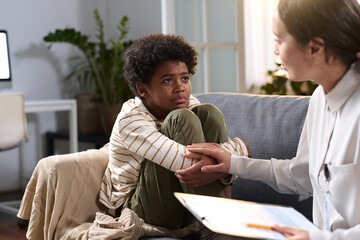 Portrait of young African American boy listening to caring psychologist in therapy session for...