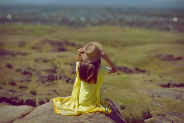 A woman in a yellow dress sits on a sandstone hilltop overlooking a vast landscape.. A woman dressed in yellow sits on a rocky hilltop, gazing out at the expanse of the land below. Her straw hat 