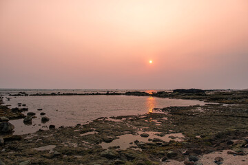 Sunset shot at the Pacific ocean shore, sun reflecting in the shallow water. Hazy colorful orange sky, blurred foreground. Los Cobanos, El Salvador.