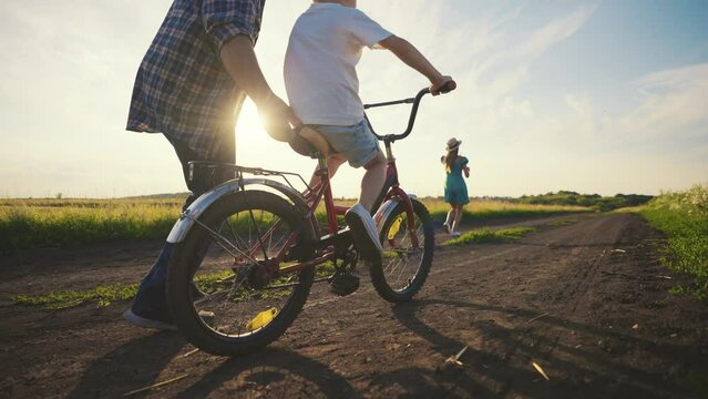 Dad helping to little son ride bike, happy family weekend in summer evening. Mother and father spending summer vacation with child in countryside, parenthood and childhood, teaching kids, parenting