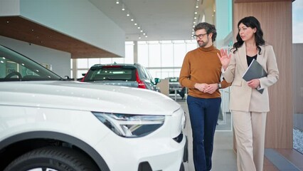 Charming Caucasian agent presenting new car model to her customer in auto salon. Pretty female manager walking with man next to while vehicle and telling about car information. Business concept. - Powered by Adobe