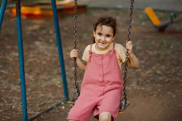 Smiling kid girl playing on a swing outdoor on a playground. Children summer activity.