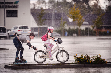 Woman on rollerblades and young girl riding a bicycle in the rain, sharing a fun and energetic outdoor moment together.