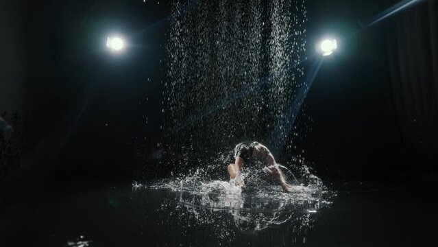 Graceful blonde woman in wet black dress sitting on floor and splashing water. Passion and emotions, beautiful slender lady playing with water in dark studio, slow motion, sexy female person moving