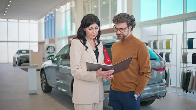 Camera view of Caucasian beautiful woman in business suit showing variants or car models in catalogue to her potential buyer. People talking together while standing in middle of auto salon.