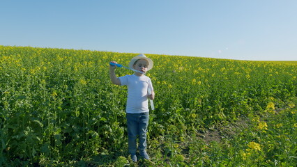 Happy Little Boy Playing In Nature. Boy Blows Soap Bubbles In Rapeseed Field. Boy Standing In Field In Summer Under Rays Of Sun And Blowing Soap Bubbles With Rainbow Reflection.