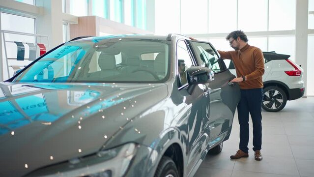 Camera view of handsome Caucasian man in glasses opening back door of car. Observing with focus new automobile in auto salon. Getting ready for buying vehicle or renting. Shopping concept.