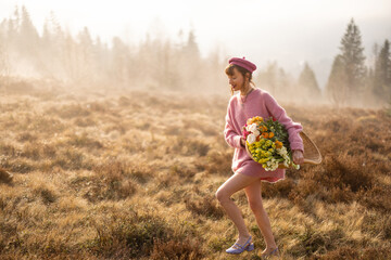 Young woman in pink walks with basket full of colorful flowers on a beautiful meadow in mountains during foggy morning. Concept of beauty and carefree lifestyle on nature
