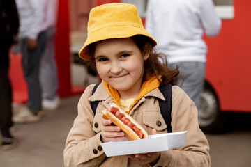 Cute little girl eating hot dog nearby food track on a street. Food delivery or take away food...