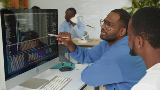 Side view of two bi-ethnic programmers sitting in front of monitor and fixing bags in script while coding at startup office