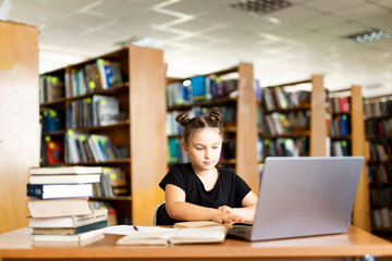 a young girl in black headphones sits at a table on which there is an open book, in a library room, studying.