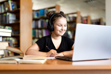School library. Schoolgirl studying online using laptop, taking notes on paper. Teenage schoolgirl...