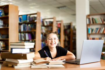 a young girl in black headphones sits at a table on which there is an open book, in a library room, studying.