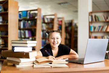 a young girl in black headphones sits at a table on which there is an open book, in a library room, studying.