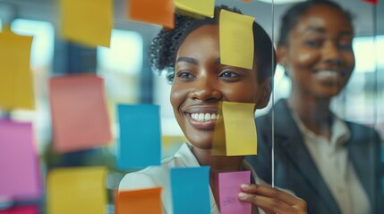 Two Black businesswomen stand smiling in a boardroom, surrounded by colorful sticky notes on a glass wall. They appear to be engaged in a collaborative planning session.