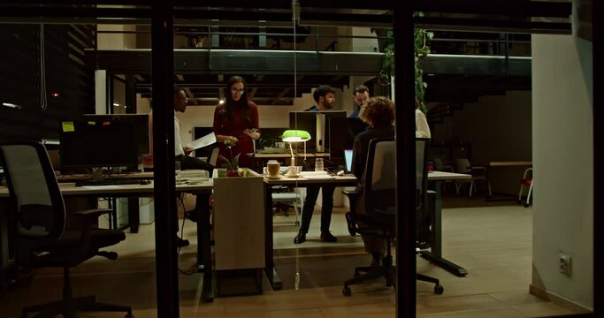 A multiethnic group of business employees collaborates on a project late at night in a contemporary office setting, illuminated by desk lamps, showcasing their commitment and teamwork.