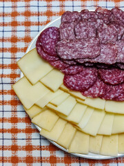 Simple feed featuring a plate adorned with two types of cheese and two types cured sausage cut into thin pieces, offering variety of flavors. Close-up on white red checkered tablecloth.