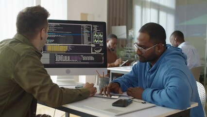 Medium shot of experienced male Caucasian programmer showing coding script on computer screen to Asian newcomer at office desk