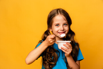Cute little brunette girl in blue t-shirt eating tasty yogurt on light yellow background. Healthy nutrition concept. Copy space.