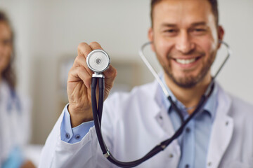 Close up portrait of a young happy smiling male doctor in white medical uniform standing with stethoscope in clinic. Confident man physician looking cheerful at camera. Selective focus.