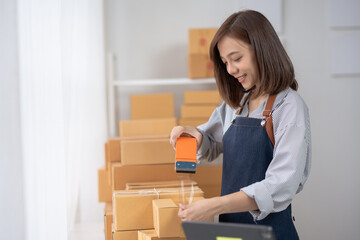 Young woman in apron packing boxes with tape dispenser in brightly lit room filled with cardboard boxes, working on online business orders. © amnaj