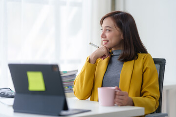 Professional woman in yellow blazer holding a coffee cup and thinking at her desk with a computer...