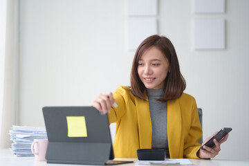 Young professional woman in a yellow blazer working on a tablet at her desk, multitasking with a...