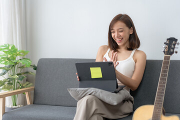 Smiling woman sitting on sofa using tablet for video call, casual indoor setting with a guitar in the background.