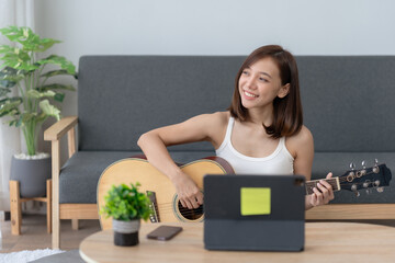 Young woman practicing guitar at home with a laptop. She is smiling and looks happy. The scene is...
