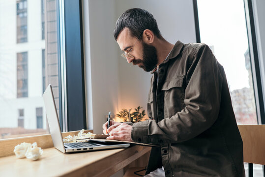 Focused writer in a cafe. Bearded man in glasses writing in a notebook near a laptop, crumpled papers around. Creative thinking process.