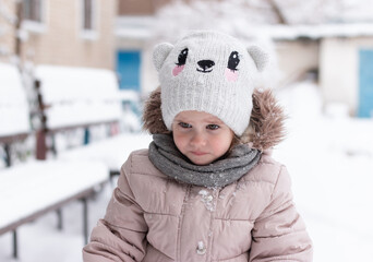 A small beautiful offended girl in a knitted hat and warm clothes outdoors in winter. Children's...