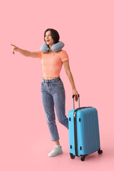 Beautiful young happy female tourist with suitcase and travel pillow pointing at something on pink background