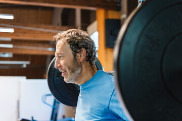 Portrait of a mature man lifts a barbell with weighted discs in a gym. He does squats.