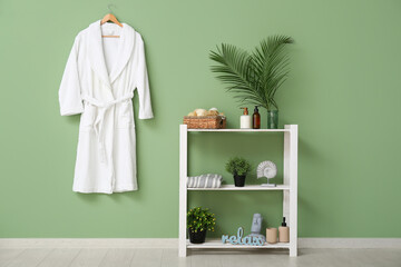 White bathrobe hanging on green wall and different bath supplies on shelves in bathroom