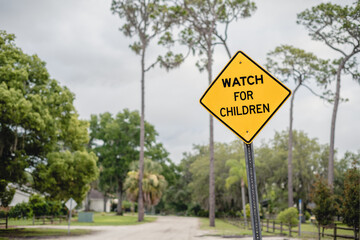Crooked WATCH FOR CHILDREN yellow, diamond sign with space on the left side and tree-lined road and fence in the background