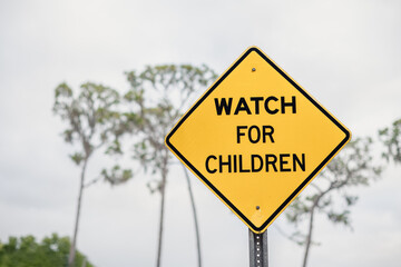 WATCH FOR CHILDREN, yellow sign, close up with sky and trees in the background on a cloudy day
