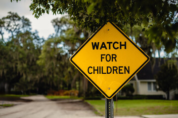 WATCH FOR CHILDREN yellow sign surrounded by foliage with a dirt road in the background, horizontal