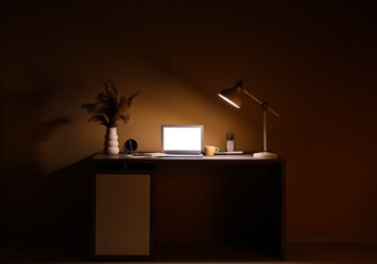 Modern workplace with glowing lamp, laptop, cup and pampas grass in evening