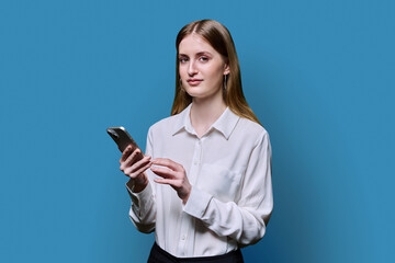 Fototapeta premium Teenage student girl in white shirt holding smartphone in hands on blue studio background