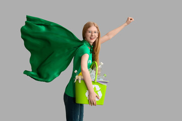 Female volunteer in superhero cape holding recycle bin with plastic on grey background