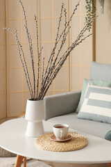 Vase with willow branches and cup of tea on table in living room