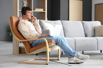 Young man with headset and laptop talking in armchair at home