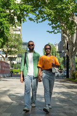 Romantic African American couple holding hands while walking down street in summer time. Serious self-confident black man and stylish girl going along road, students spend free time together strolling