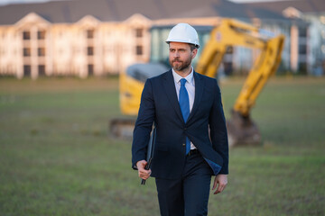 Construction manager in suit and helmet at a construction site. Construction manager worker or supervisor wearing hardhat in front of house. Supervisor construction manager near excavator. Renovation.