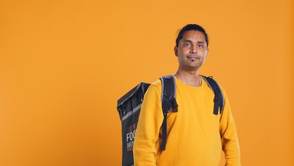 Portrait of confident indian man holding thermal backpack, crossing arms, studio background. Radiant nonchalant delivery person prepared to deliver takeaway food to customers, camera B