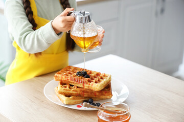 Cute little girl pouring honey on waffles in kitchen, closeup