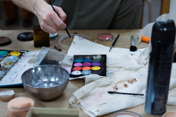 Obraz premium Messy colorful creative table with makeup palette, paints, brushes, pencil sharpener, paintbrushes set and bowl of water, paper tissues on wooden table. Stage make up. Creative chaos. Selective focus