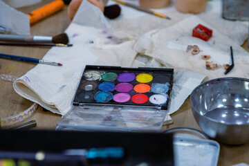 Messy colorful creative table with makeup palette, paints, brushes, pencil sharpener, paintbrushes set and bowl of water, paper tissues on wooden table. Stage make up. Creative chaos. Selective focus