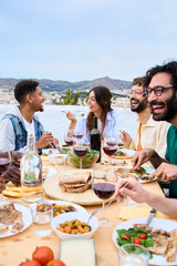 Vertical. Happy people around meal table with wine glasses and food celebration terrace barbecue party. Young multiracial friends enjoying laughing together lunch time on summer weekend on rooftop