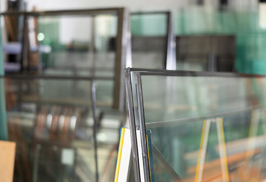 Close-up of insulated glass units stacked on support stand in workshop, with various glass panels and industrial equipment in blurred background